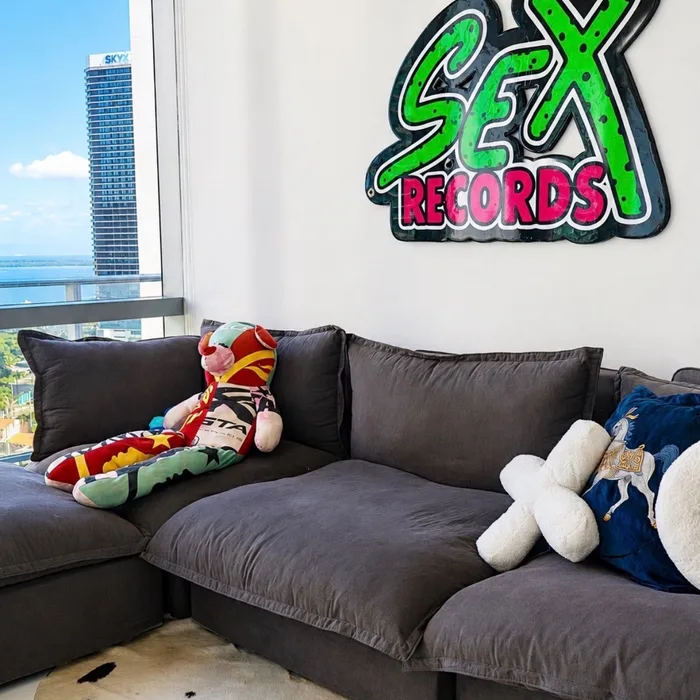 A glossy green and pink SEX RECORDS sign mounted above a grey sectional in a Brickell high-rise, with the bay through the window